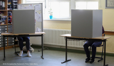 Personen sitzen einzeln hinter einem geschützten Wahltisch