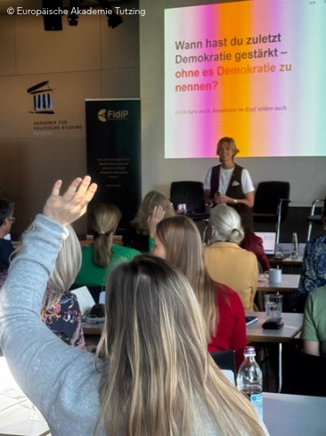 Kurzhaarige Frau steht vor einer großen Bildfläche mit einer bunten Präsentation - vor ihr sitzt ein Publikum aus Frauen, die sich durch Handmeldungen aktiv beteiligen.